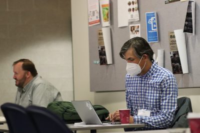 Roger Travis (wearing a white and blue checkered collared shirt) gazes down at a laptop while seated in the basement of the ITE building