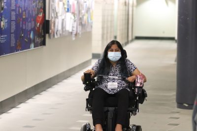 Arpita Kurdekar (seated in electric wheelchair wearing black pants and a blue/white-flower patterned shirt) smiles at the camera in the basement of the ITE building