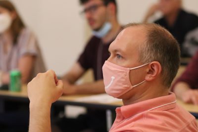 Graham Benedict (wearing a pink polo shirt with pink mask featuring a white smile) watches a presentation with his left elbow on the desk in front of him, arm pointed up with a fist; other attendees visible in background (blurred)