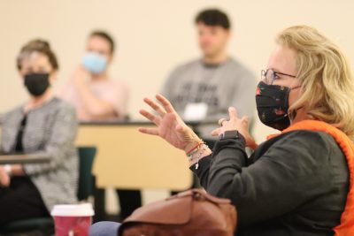 Manuela Wagner (black jacket, orange scarf, brown glasses, black mask) gestures while asking a question during Wendi Sierra's presentation (Clarissa Ceglio, Joshua Hirshfield, and James Coltrain are visible in the background [blurred])