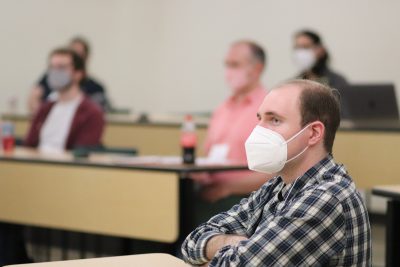 Landon Bassett (wearing a white, green, and black plaid collared shirt) watches a presentation in an ITE classroom; other Frontiers in Playful Learning attendees are visible in the background (blurred)