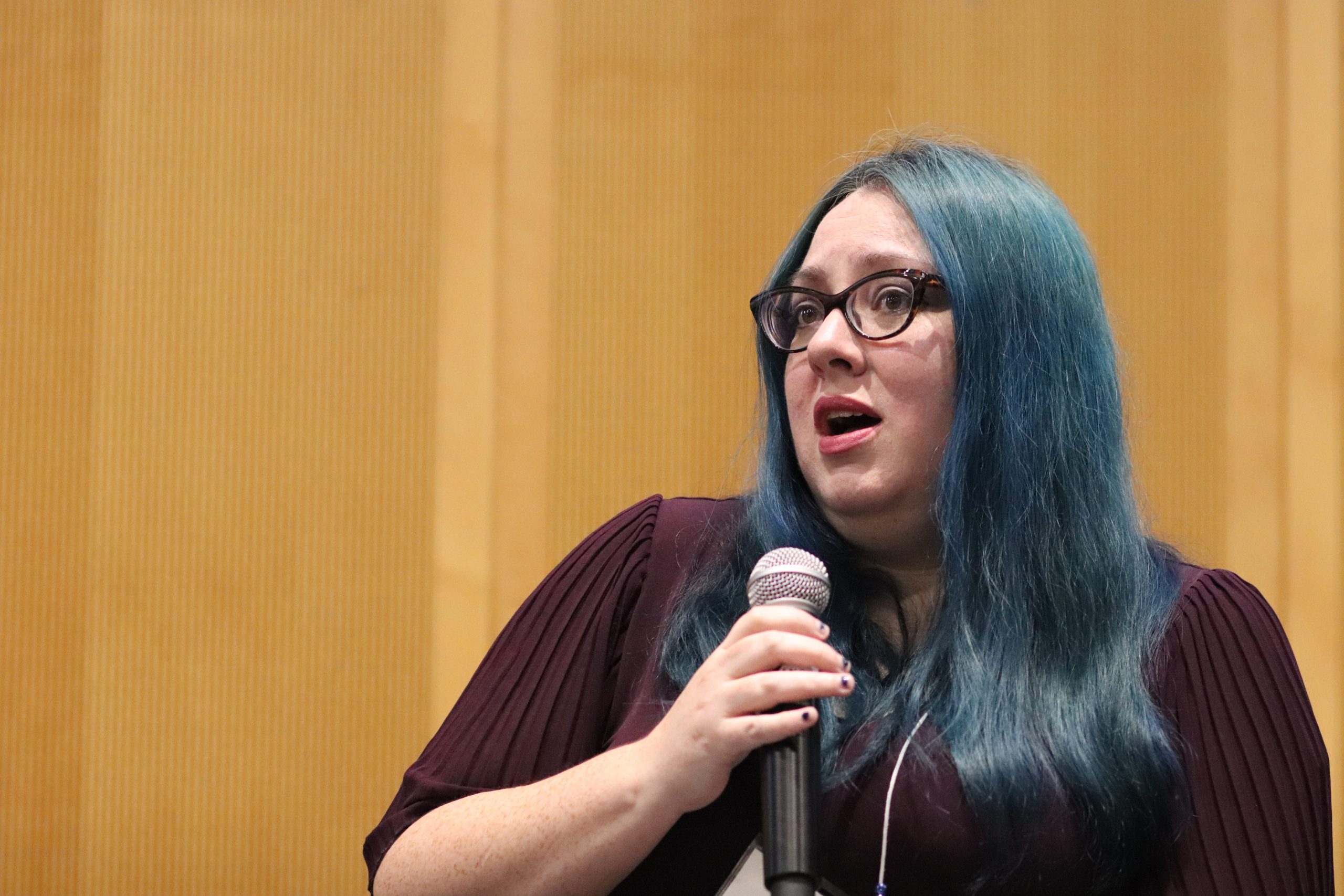 Wendi Sierra (blue hair, dark rimmed glasses, purple sweater) speaks on the ITE C80 stage during a panel discussion about the humanities