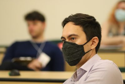 Tom Lee (wearing a light collared shirt and black mask) looks on during a presentation in an ITE classroom; James Coltrain is visible (blurred) in the background