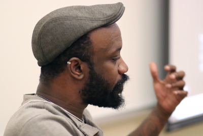 Drew John Ladd speaks to an audience, holding left hand upward with palm facing himself (wearing a gray shirt and dark grey cap)