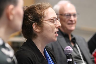 Jessica Creane speaks with a microphone in hand during a Frontiers in Playful Learning panel discussion; Michael Young is pictured in background (blurred); Caro Murphy is pictured in the foreground (blurred)