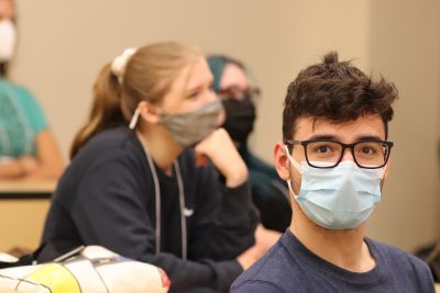 Jake Tomassi (blue t-shirt and black glasses) looks at the camera during a presentation in an ITE classroom; Mackenzie Fox, Wendi Sierra, and Tori Wagner are visible in the background (blurred)