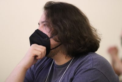 Colter Moos (wearing a blue t-shirt and black mask) grasps his chin with his right hand while listening intently to a presentation